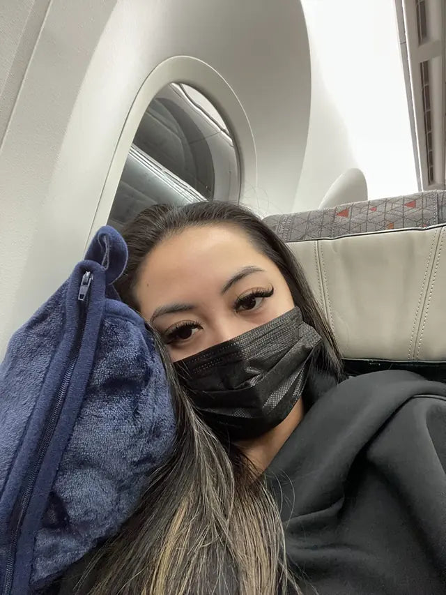 A passenger takes a selfie beside an airplane window while resting against a navy EverSnug travel pillow